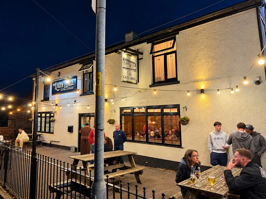 Lazy Hill Tavern in Aldridge at dusk, a cream-painted pub with festoon lights and people at picnic benches outside