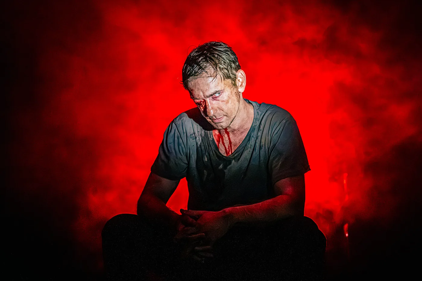 A performer in torn clothing with prosthetic wounds, head bowed, lit red through smoke