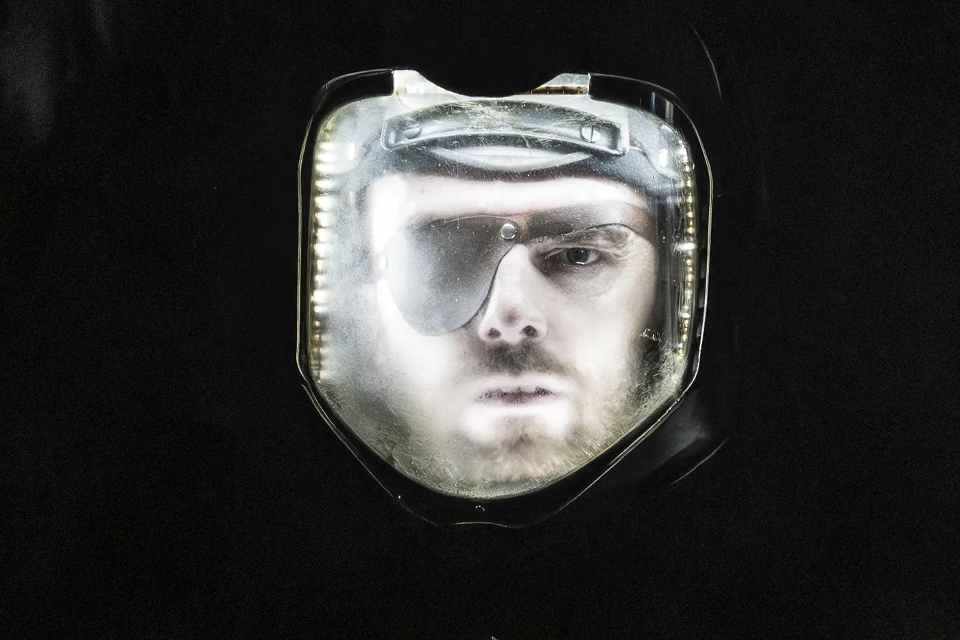 A face lit from below inside a scratched glass space helmet, surrounded by darkness