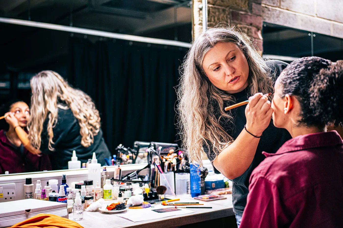 Two makeup artists working on performers at a mirror lined with brushes and pots
